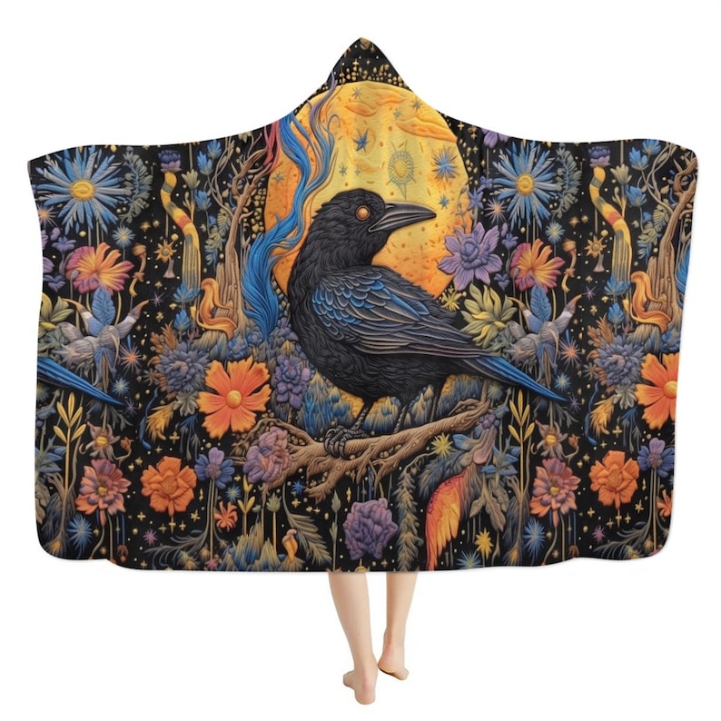 May include: A hooded cloak with a detailed illustration of a black raven perched on a branch, set against a full moon and colourful flowers. The cloak has a black background with vibrant floral and celestial elements.