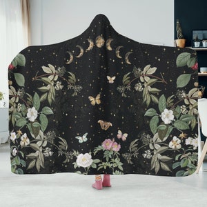 May include: Black hooded blanket with a floral and moon pattern. The pattern features white flowers, green leaves, butterflies, and gold moons on a black background.