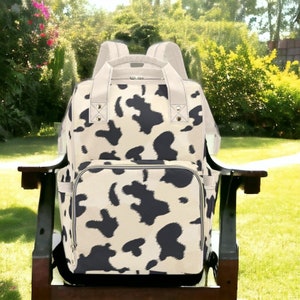 K&ouml;nnte beinhalten: Ein schwarzer und wei&szlig;er Kuhflecken-Wickeltasche-Rucksack mit einer Rei&szlig;verschlusstasche vorne und verstellbaren Tr&auml;gern.