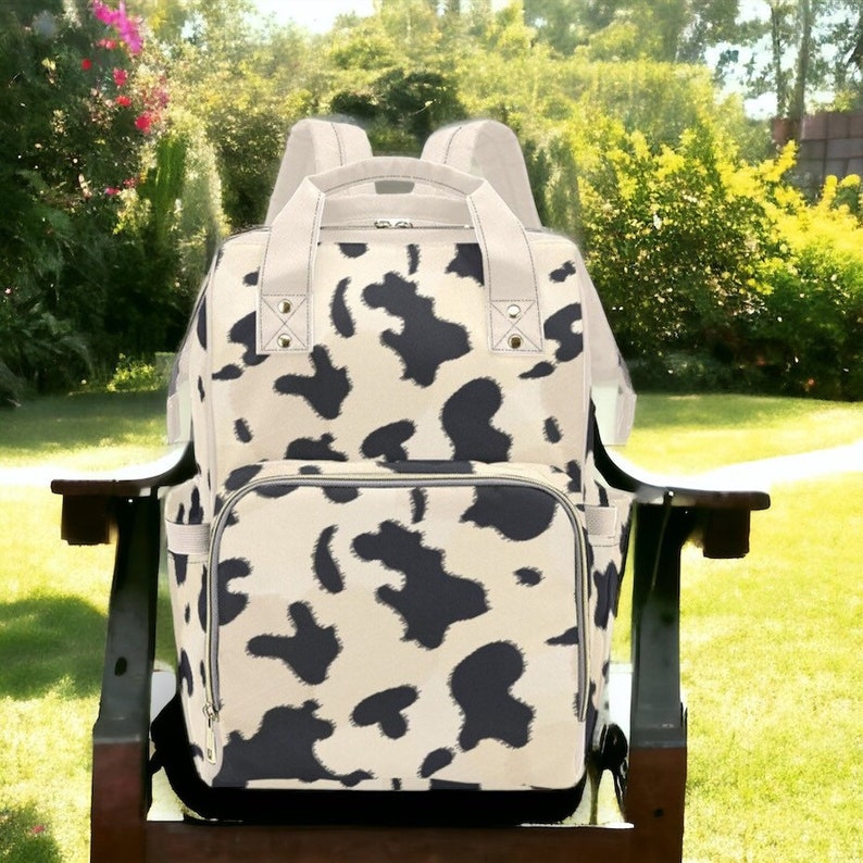 K&ouml;nnte beinhalten: Ein schwarzer und wei&szlig;er Kuhflecken-Wickeltasche-Rucksack mit einer Rei&szlig;verschlusstasche vorne und verstellbaren Tr&auml;gern.