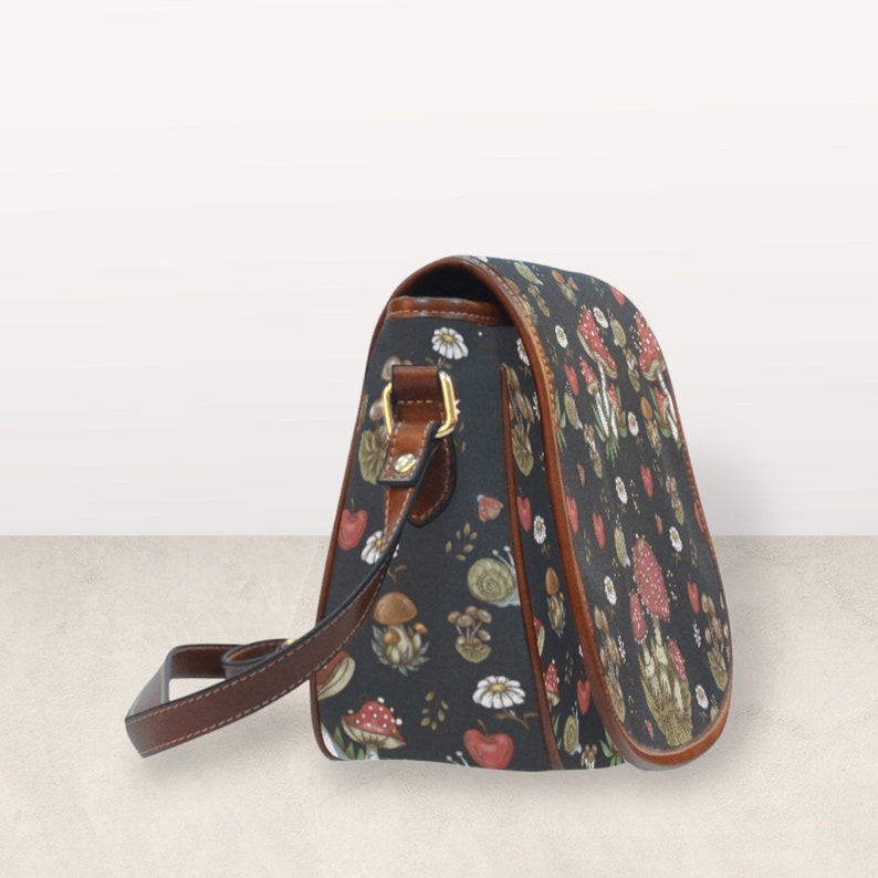 May include: A black crossbody bag with a brown leather strap and a mushroom and flower print. The bag has a flap closure and a gold buckle.