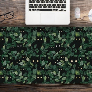 May include: A black desk mat featuring a repeating pattern of black cats peeking through green leaves. The cats have yellow eyes, and the leaves are various shades of green. The mat is rectangular and sits on a wooden desk.