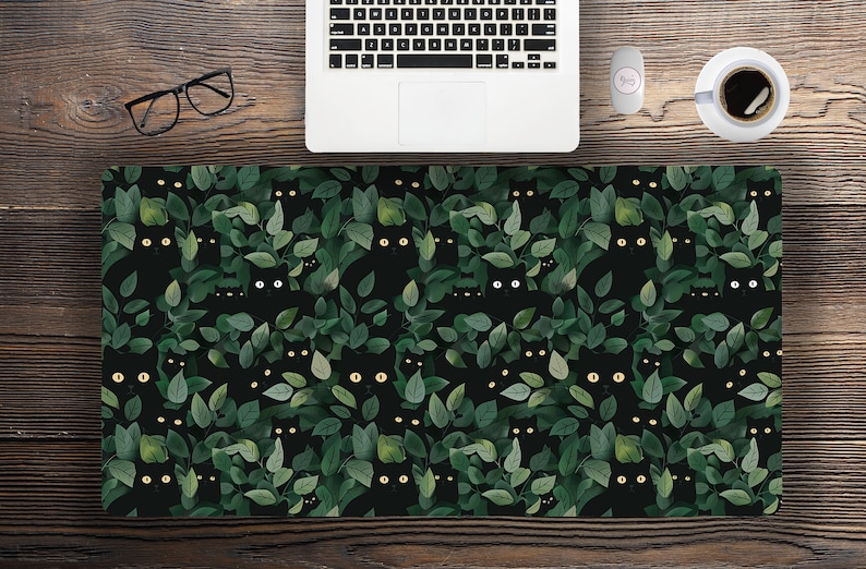 May include: A black desk mat featuring a repeating pattern of black cats peeking through green leaves. The cats have yellow eyes, and the leaves are various shades of green. The mat is rectangular and sits on a wooden desk.