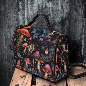 May include: A black satchel handbag with a mushroom and floral print. The bag has a top flap closure and a gold-toned buckle. The bag has a long adjustable strap.