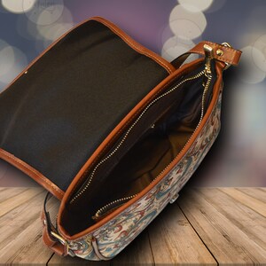 May include: Open, patterned shoulder bag with a brown leather trim and strap. The bag features a gold zipper and a black interior. The exterior has a colorful floral design with red, blue, and cream colors.