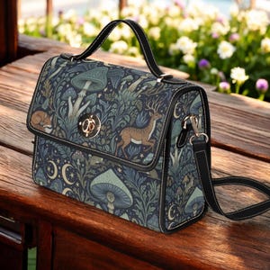 Può includere: Borsa a tracolla blu scuro con un motivo forestale stravagante con funghi, cervi e volpi. La borsa ha un manico e una tracolla neri, con una chiusura color oro. La borsa è appoggiata su una superficie di legno.