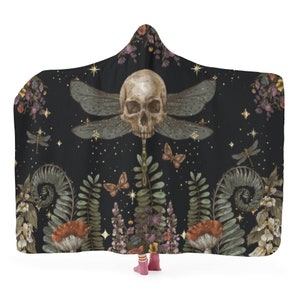 May include: Black hooded blanket with a skull and dragonfly wing design. The blanket features a detailed illustration of a skull with dragonfly wings, butterflies, and botanical elements. The color palette includes black, beige, green, and purple.