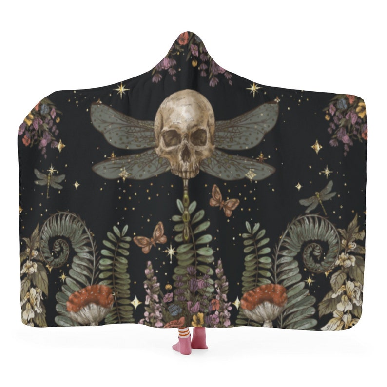 May include: Black hooded blanket with a skull and dragonfly wing design. The blanket features a detailed illustration of a skull with dragonfly wings, butterflies, and botanical elements. The color palette includes black, beige, green, and purple.