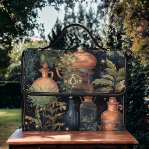 May include: A black handbag with a botanical illustration featuring copper-toned vessels and lush greenery. The bag has a black handle, a gold clasp, and a white stitched border. The background is a blurred outdoor scene.