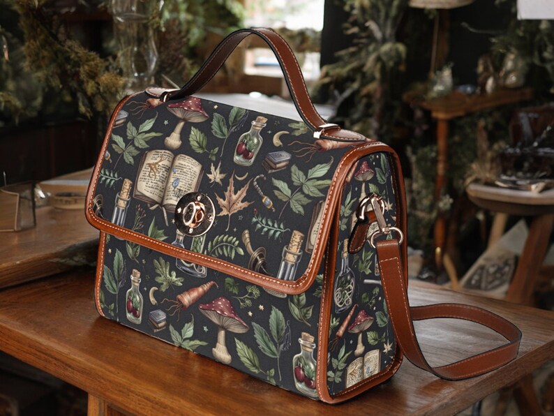 May include: A brown leather satchel with a black canvas body featuring a whimsical pattern of mushrooms, leaves, and potion bottles. The bag has a top handle and a long adjustable shoulder strap.