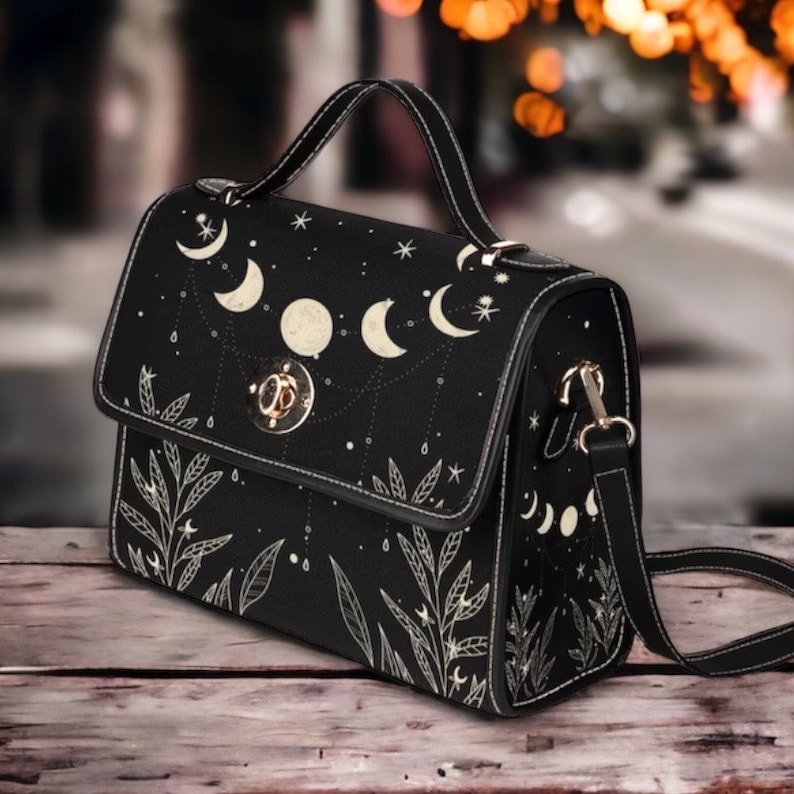 May include: A black crossbody bag with a white moon and star design. The bag has a top handle and a long adjustable strap.
