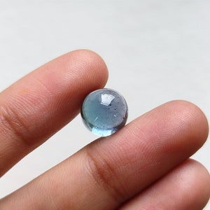 May include: A clear, round, blue-green glass marble. The marble is held between two fingers, showcasing its translucent quality and subtle color variations. The marble's surface reflects light, creating highlights.