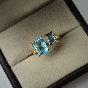 Könnte beinhalten: Ein goldener Ring mit zwei blauen Edelsteinen. Der größere Stein ist hellblau, während der kleinere Stein dunkelblau ist.