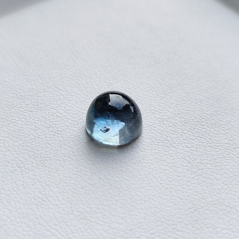May include: A cabochon gemstone with a rounded top, displaying a gradient of colors from deep blue to light blue. The polished surface reflects light, creating a glossy appearance. The gemstone is set against a white background.