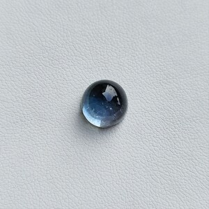 May include: A round, polished gemstone with a gradient of blue hues. The gem transitions from a lighter, translucent blue at the base to a darker, opaque blue at the top. The surface reflects light, creating a glossy appearance.