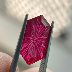 May include: A vibrant, deep pink, faceted gemstone with a complex, star-like geometric pattern cut into its surface. The stone is held securely by a pair of metal tweezers, showcasing its translucent quality and sharp, angular edges.