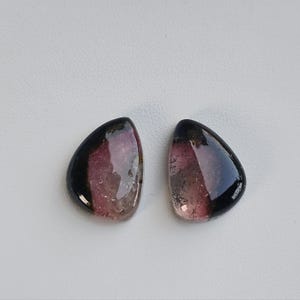 May include: Two polished, teardrop-shaped gemstones. Each stone features a gradient of colors, transitioning from black to a deep pink and a translucent base. The stones are set against a white background.