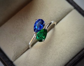 Pode incluir: Um anel de prata com uma gema azul em forma de pêra e uma gema verde em forma de pêra. O anel é apresentado numa caixa de anel de veludo.
