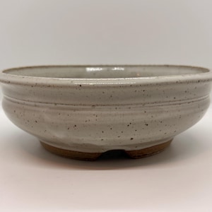 May include: A speckled, off-white ceramic bowl with a wide, shallow design. The bowl has a rounded shape with a slightly flared rim and a textured surface. The base is a darker brown color. This bowl is suitable for plants or decorative purposes.