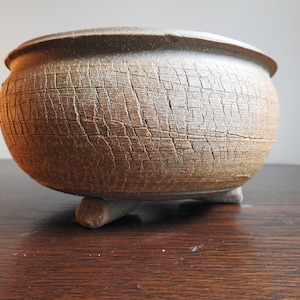May include: A handcrafted, round, brown ceramic planter with a textured, cracked surface. The planter rests on three small, rounded feet. The rim of the planter is smooth and dark brown. A decorative piece for plants.