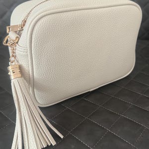 May include: An off-white crossbody bag with a textured finish. It has a gold-coloured zip and hardware, plus a matching tassel. The bag is rectangular with rounded corners and a structured shape.