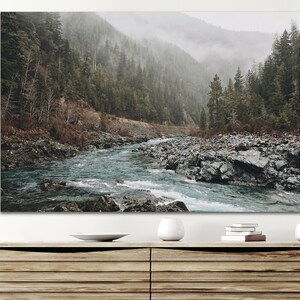 May include: A misty forest scene with a river flowing through the center of the image. The river is surrounded by rocks and the forest is dense with trees. The sky is overcast and the air is thick with fog.