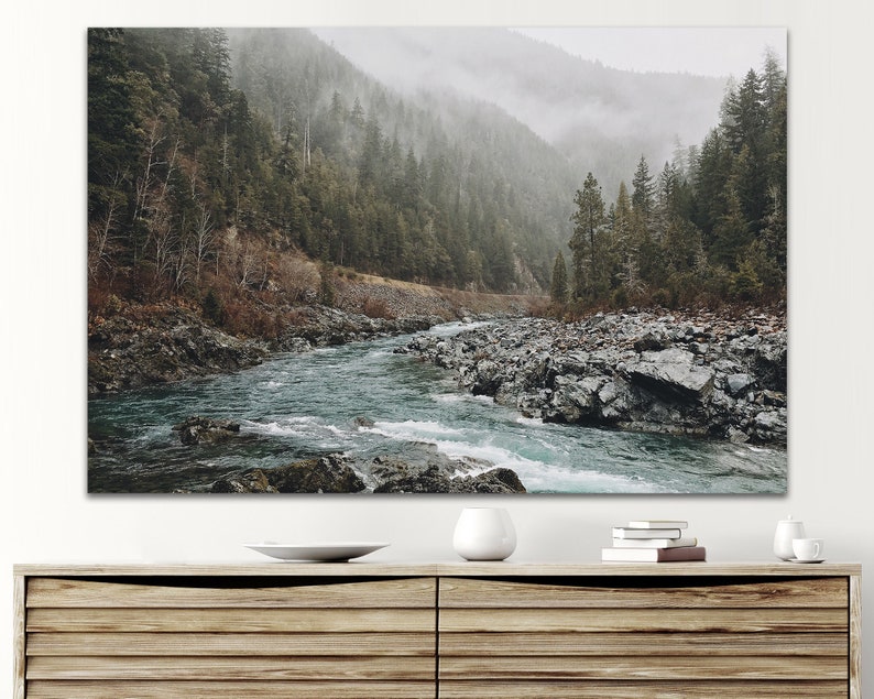 May include: A misty forest scene with a river flowing through the center of the image. The river is surrounded by rocks and the forest is dense with trees. The sky is overcast and the air is thick with fog.