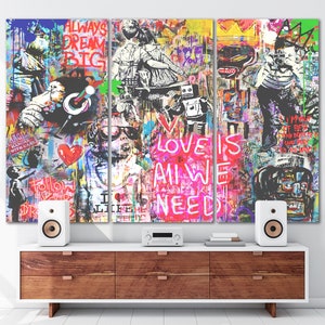 May include: A colourful triptych canvas print with a graffiti style design. The centre panel features the words "Love is all we need" in pink and white lettering. The left panel features a figure wearing a red heart and the right panel features a figure holding a camera.