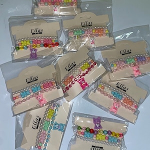 Puede incluir: Múltiples pulseras con cuentas coloridas y pequeños dijes de ositos de peluche se exhiben en envases de plástico transparente. Las pulseras presentan una mezcla de colores pastel y vibrantes, incluyendo rosa, amarillo, azul y verde. Cada paquete tiene una etiqueta que dice "Ellas".