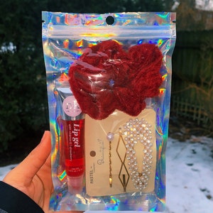 May include: A clear, iridescent package containing a red lip gel, two red velvet scrunchies, and two hair accessories. One hair accessory is a gold bobby pin with a pearl, and the other is a pearl and rhinestone hair clip.