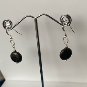 Può includere: Un paio di orecchini pendenti in argento con pietre ovali nere. Gli orecchini sono appesi a un supporto in argento.