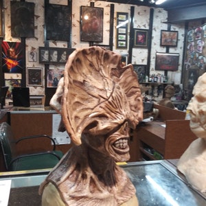 May include: A detailed, brown, sculpted bust of a creature with a large, fleshy head and a gaping mouth. The creature has a textured, wrinkled surface and appears to be made of a rubbery material. The bust is displayed on a glass surface in a shop setting.