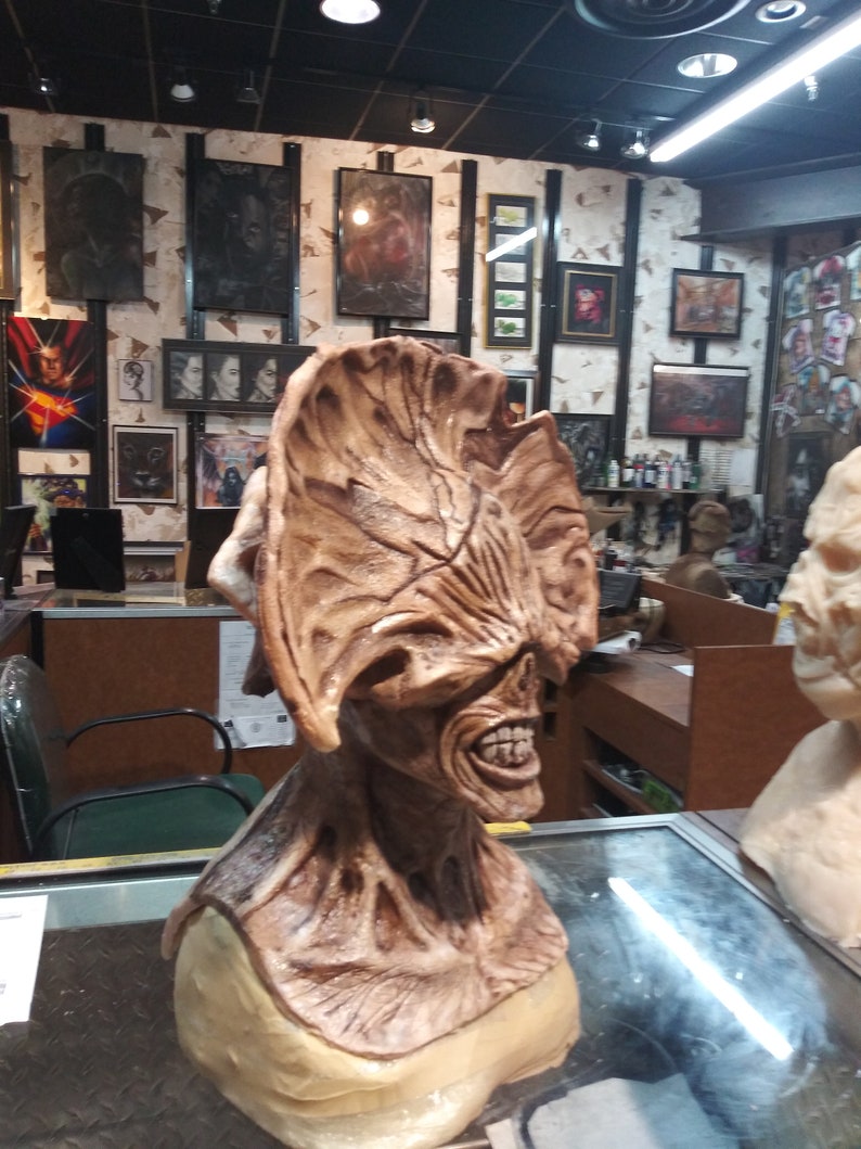 May include: A detailed, brown, sculpted bust of a creature with a large, fleshy head and a gaping mouth. The creature has a textured, wrinkled surface and appears to be made of a rubbery material. The bust is displayed on a glass surface in a shop setting.