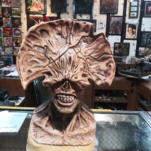 May include: A brown and beige sculpted bust of a monster with a large, open mouth and a toothy grin. The monster has a large, fleshy head with a series of ridges and folds. The bust is displayed on a glass surface.