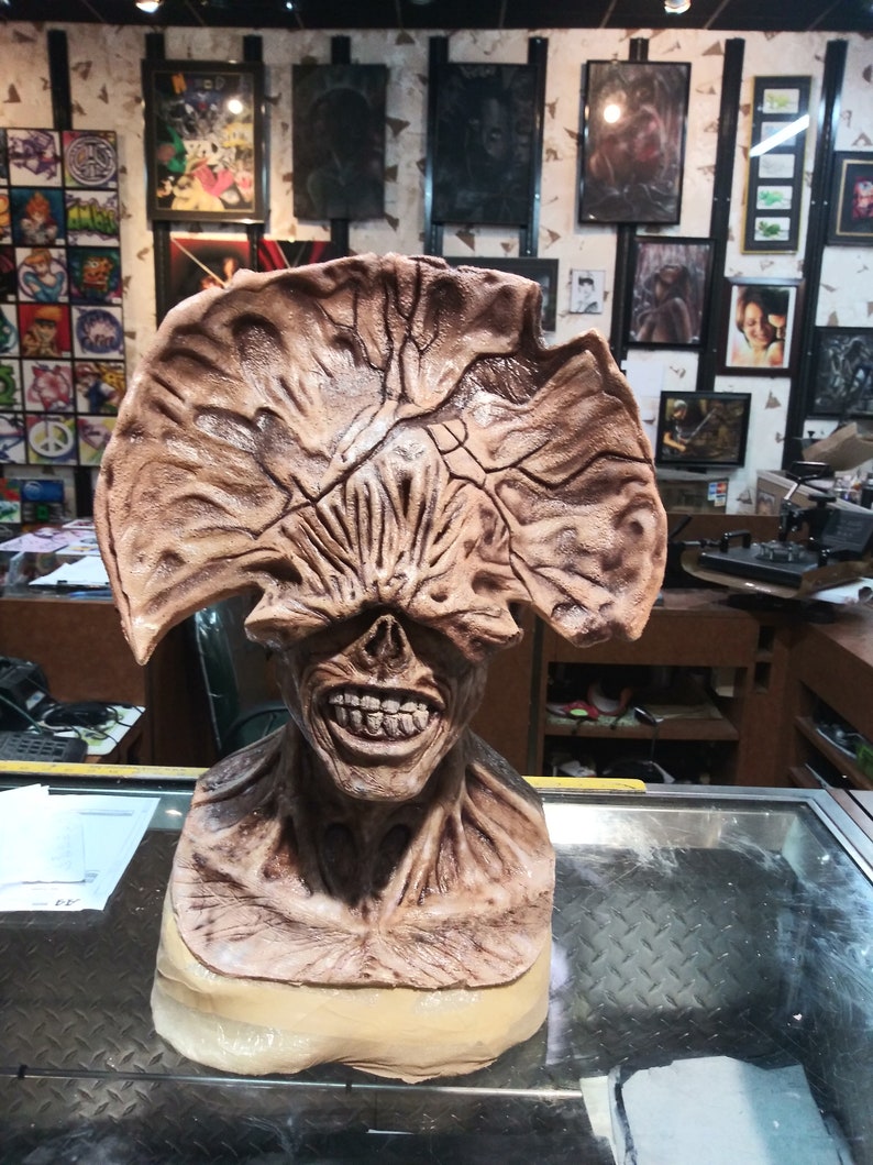 May include: A brown and beige sculpted bust of a monster with a large, open mouth and a toothy grin. The monster has a large, fleshy head with a series of ridges and folds. The bust is displayed on a glass surface.