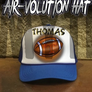 May include: A white and blue trucker hat with a blue brim. The hat has a white mesh back and a white front panel with a brown football and the name "THOMAS" printed in black.
