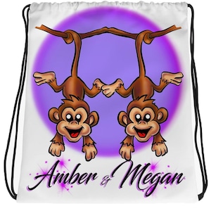 May include: White drawstring bag with a purple circle and two brown monkeys hanging upside down from a branch. The monkeys are smiling and have big eyes. The text "Amber & Megan" is written below the monkeys in purple.