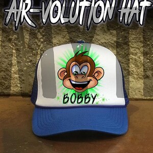 May include: A blue and white trucker hat with a cartoon monkey design and the name "BOBBY" written in green. The hat is part of the "AIR-VOLUTION HAT" collection.