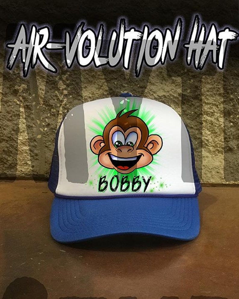 May include: A blue and white trucker hat with a cartoon monkey design and the name "BOBBY" written in green. The hat is part of the "AIR-VOLUTION HAT" collection.