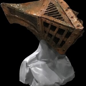 May include: A weathered, metallic-looking helmet with a cage-like design and riveted details, mounted on a gray bust. The helmet has a rusty brown and bronze color scheme, with a dark background.
