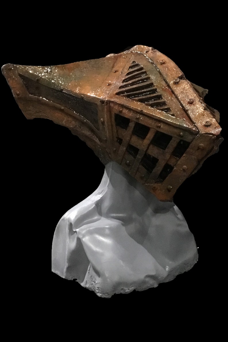 May include: A weathered, metallic-looking helmet with a cage-like design and riveted details, mounted on a gray bust. The helmet has a rusty brown and bronze color scheme, with a dark background.