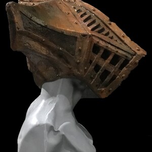 May include: A sculpture of a head with a cage-like structure on top. The cage is made of dark brown metal with rivets and a grid pattern. The head is a light gray color, and the background is black.