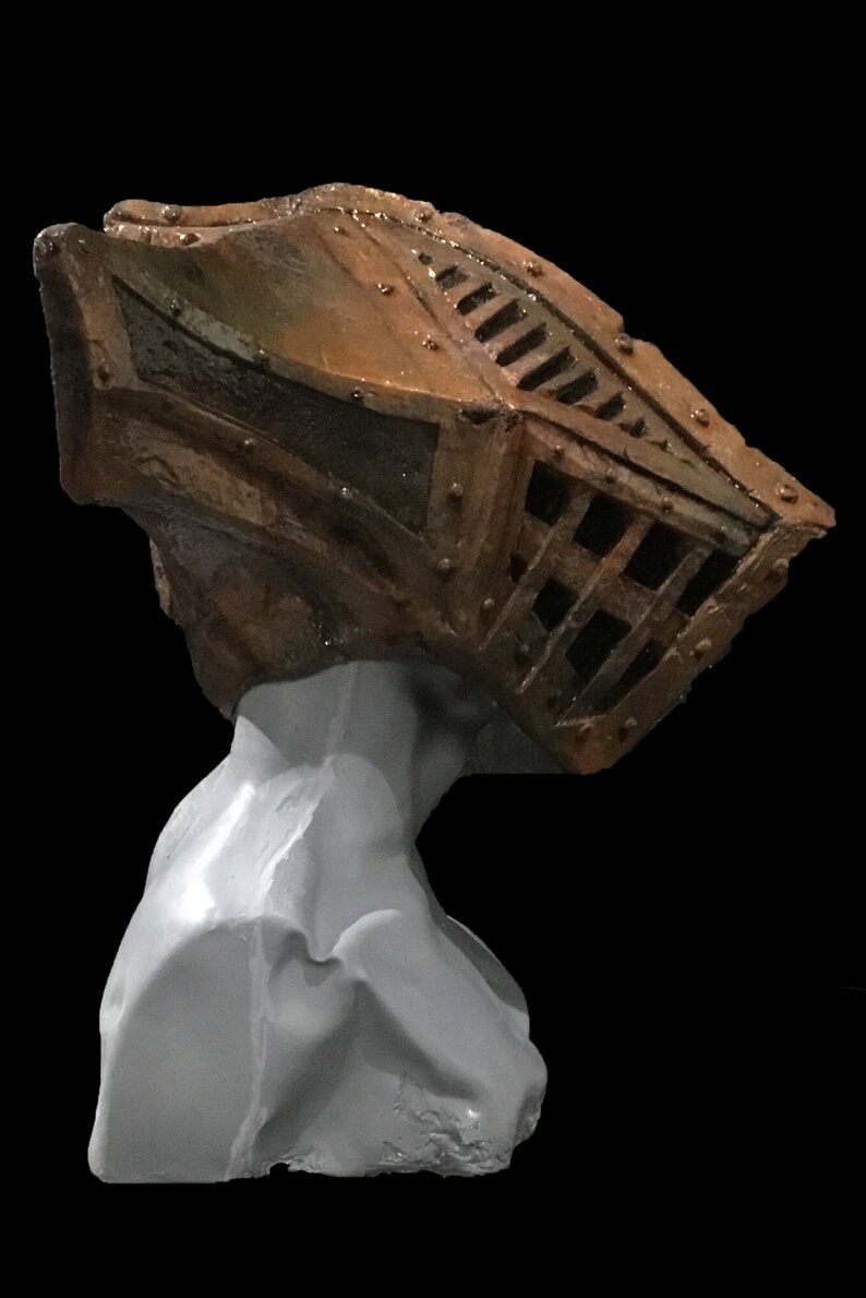 May include: A sculpture of a head with a cage-like structure on top. The cage is made of dark brown metal with rivets and a grid pattern. The head is a light gray color, and the background is black.