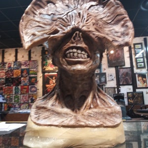 May include: A brown and beige sculpture of a monster with a large, fleshy, open mouth and a closed eye. The monster's head is tilted slightly to the right. The sculpture is on a display stand.