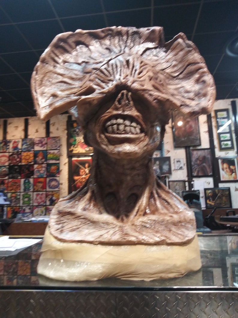 May include: A brown and beige sculpture of a monster with a large, fleshy, open mouth and a closed eye. The monster's head is tilted slightly to the right. The sculpture is on a display stand.