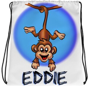 May include: White drawstring bag with a cartoon monkey hanging upside down from a branch. The monkey has brown fur, a blue circle around it, and a blue background. The bag has the name "EDDIE" written in black at the bottom.