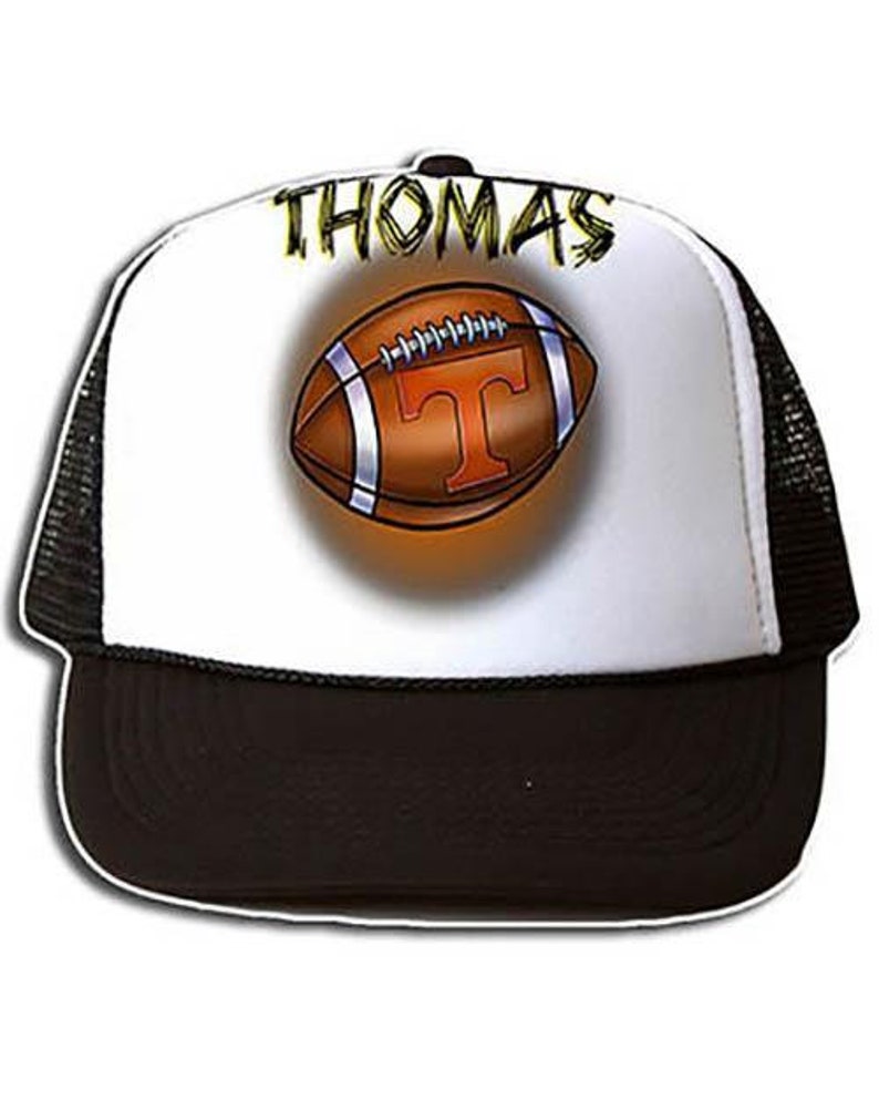May include: A white and black trucker hat with a black mesh back. The front of the hat features a brown football with a white 'T' and the name 'THOMAS' in yellow lettering.