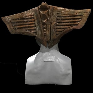 May include: A bronze-colored, stylized headpiece with a wing-like design, set against a black background. The headpiece is mounted on a gray bust, showcasing intricate details and textures. The overall aesthetic suggests a historical or fantasy theme.