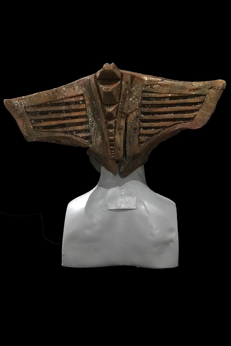 May include: A bronze-colored, stylized headpiece with a wing-like design, set against a black background. The headpiece is mounted on a gray bust, showcasing intricate details and textures. The overall aesthetic suggests a historical or fantasy theme.