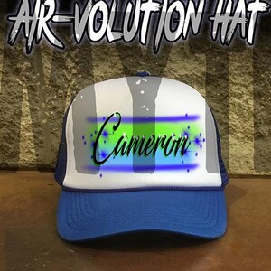 May include: A blue and white trucker hat with a mesh back. The hat has a green and blue airbrushed design with the name "Cameron" written in black.